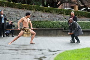 Manele crouches down as he faces a bare-chested Māori warrior, in traditional skirt, who is holding a spear