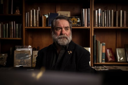 Tim White with full beard wears a black blazer and black collared shirt sits in front of shelves of books