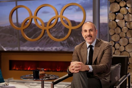 He sits in a chair in a studio, smiling, with the Olympic logo in the background
