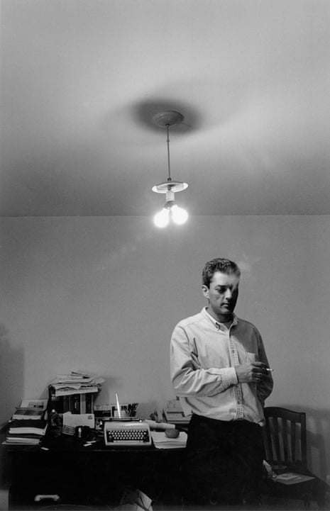 A monochrome image of Paul Auster standing in his study with a cigarette in his hand and a number of objects on a table behind him, including a manual typewriter.