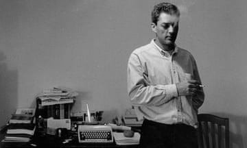 A monochrome image of Paul Auster standing in his study with a cigarette in his hand and a number of objects on a table behind him, including a manual typewriter