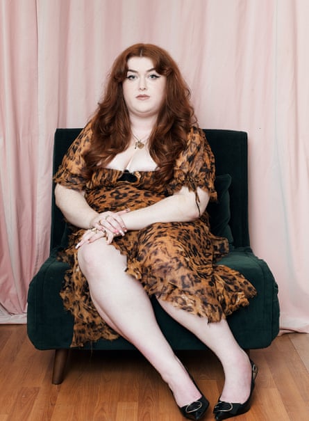 Ione Gamble sitting on a dark-green velvet chair in front of a pair of pale-pink curtains in her flat, wearing a black and brown leopard-skin patterned dress and black shoes, with both hands on her right knee, looking at the camera