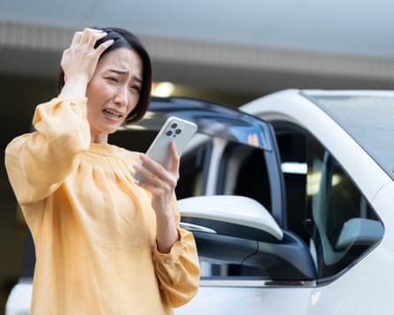 An asian women in a yellow blouse holds a hand to her head and looks distressed as she stands beside her car.
