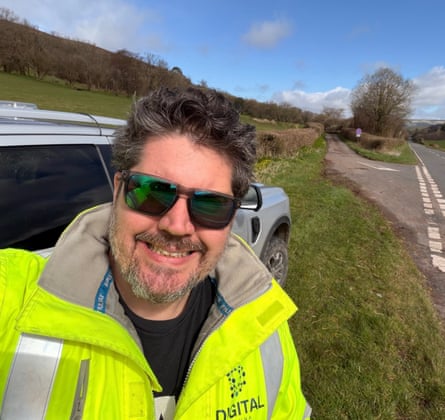 A man wearing sunglasses and a hi-vis jacket smiles as he stands on the side of the road.