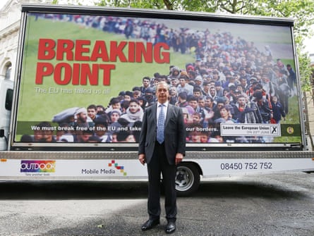A man in a grey suit and a blue tie standing in front of a big poster with the words ‘breaking point’ and lots of migrants in the background.