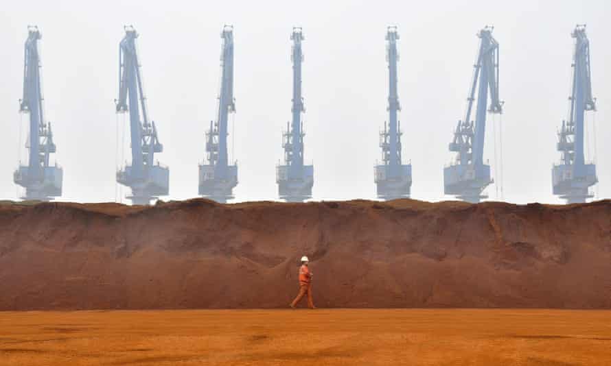 A worker walks past huge piles of red-orange iron ore