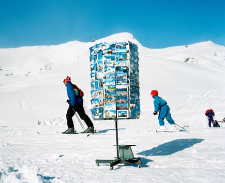 Skiers against a white mountain and blue sky, with a postcard rack standing in front of them, in 1990