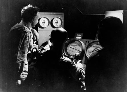 Three men stand and sit in an aircraft cockpit looking at large dials for a countdown.