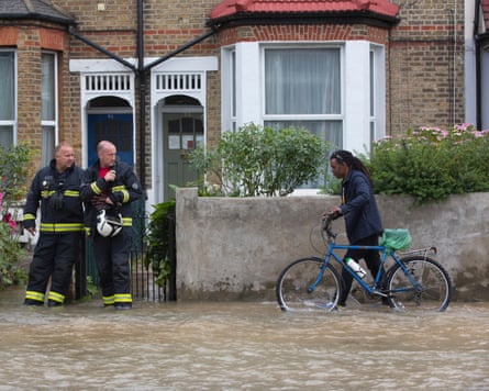 A man pushes his bike through flood water on a residential street as two firefighters look on