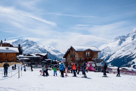 A row of skiers of all ages in a snowy village with wooden chalets and mountain peaks behind