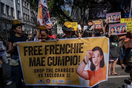 People holding banners and wearing signs on ther hats which read Free Frenchie Mae Cumpio stand in front of railings which surround a building.