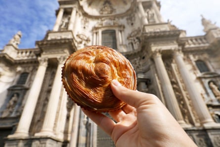 A hand holding up a pastel de carne in front of Murcia Cathedral