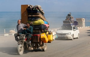 Two men ride at the front of a three-wheeled motorcycle piled high with belongings on a coastal road