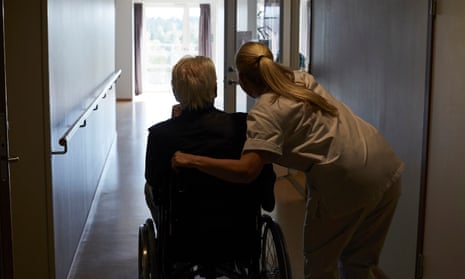 Rear view of female nurse wearing white with senior man sitting in wheelchair in a hospital corridor. She is leaning down to him with her hand on the back of his chair and they are looking down the corridor