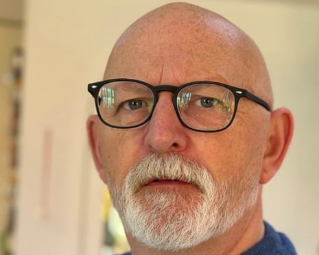 Shaun Burton, a bald man with black-framed glasses and a white beard, looks directly at the camera