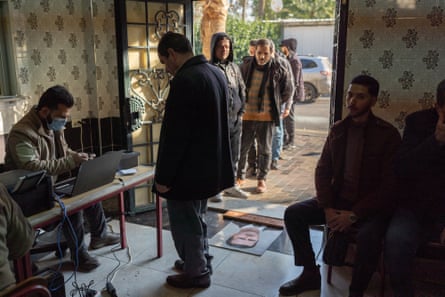 Men queue at a settlement centre where a man wearing a protective mask sits with a laptop behind a desk. A portrait of Bashar al-Assad is used as a doormat