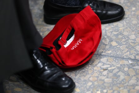 A red Maha baseball cap on the ground in between two black wingtip shoes.