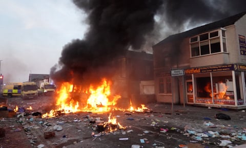 A burning police van, with three other police vans parked behind it, in a street strewn with debris