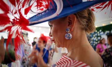 A woman wearing an extravagant blue hat with red and white feathers, large blue and white earrings and a red and white striped dress