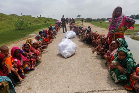 South Asian women, some with children, squat in two queues by a river with soldiers standing guard