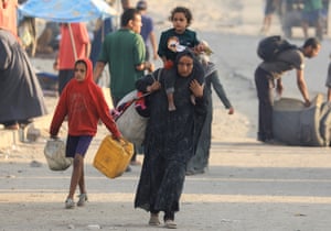 A woman in a chador with a large bag of belongings and a child on her shoulders walks down a street with another child carrying a water container beside her