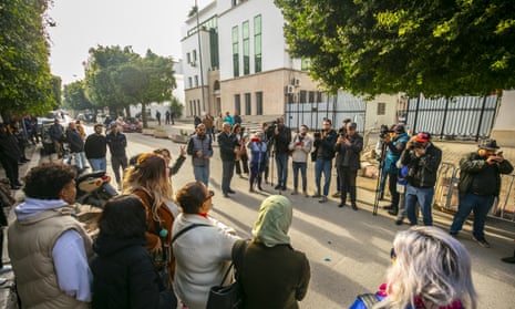 A group of people stand in the street opposite a court building, with a line of photographers and camera crew facing them on the other side of the road