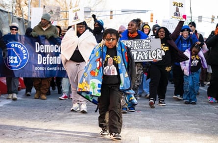 A child walks with a blanket around her shoulders in front of a group of demonstrators marching with signs