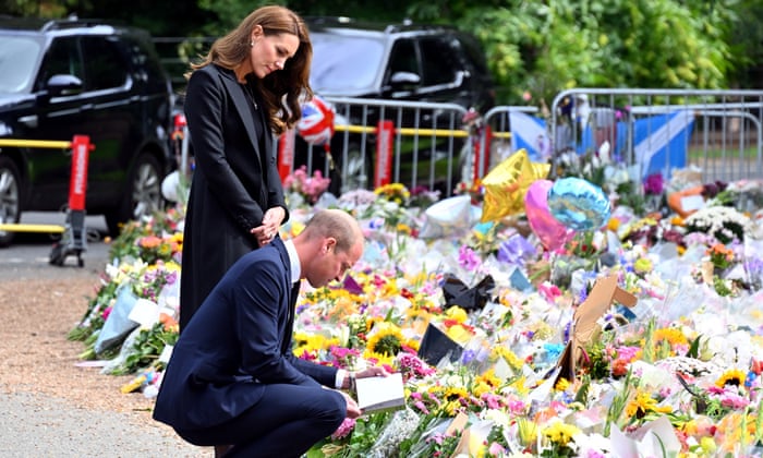 The Prince of Wales crouches while reading a card left with floral tributes to the Queen. The Prince of Wales stands, reading the card over his shoulder. 