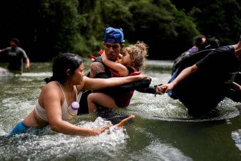 A man carries a child through a river while a woman wades beside them