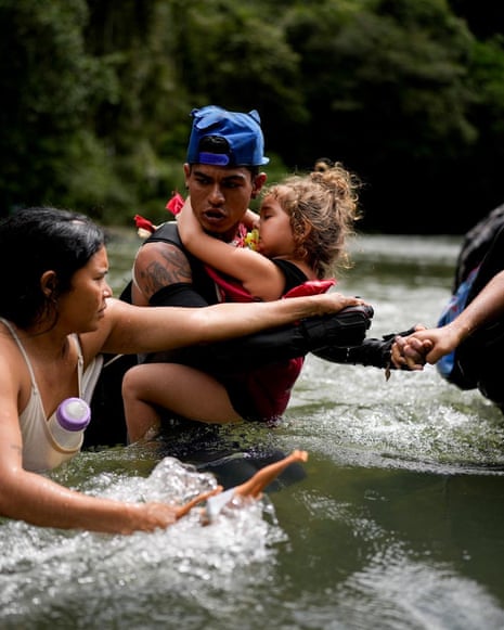 A man carries a child through a river while a woman wades beside them