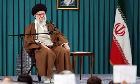 Iran’s leader, a man, sits in a chair on a stage next to the Iranian flag 