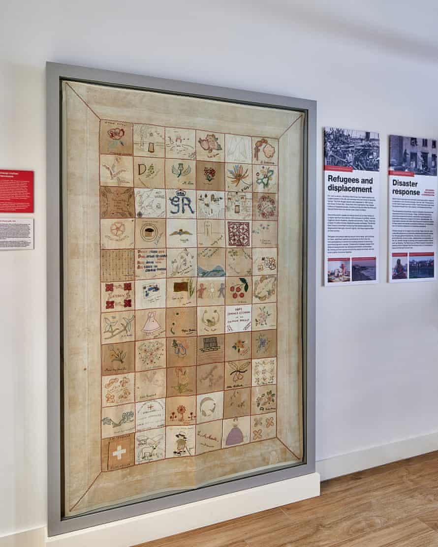 A large quilt of many squares hanging on the wall of a museum in a large glass-fronted frame