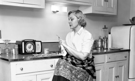 Gwyn Steinbeck in 1957, wearing a button-down shirt and a skirt, sitting near a kitchen counter with a radio and cupboards in her home
