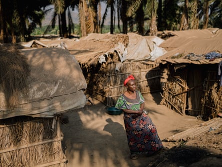 A woman in a bright red headband stands among makeshift straw houses.