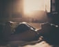 A sad woman lies on her back in bed, eyes closed, sunlight streaming through a window behind her