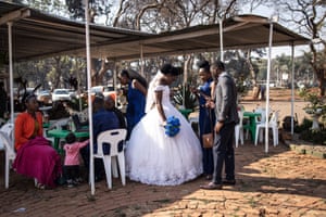 A bride stands holding a blue bouquet flanked by a small group standing and seated at tables under a corrugated iron awning