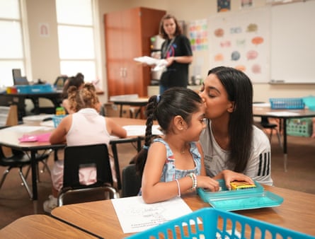 A mother kisses her daughter on the forehead in a classroom as a teacher looks on