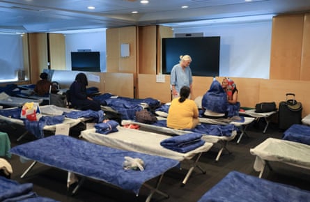 Rist speaks to woman sitting on camp-beds set up in a room where the blinds have been drawn