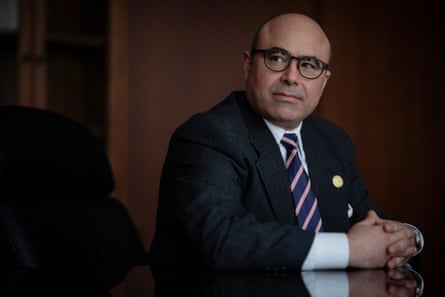 A man in a suit and glasses, sitting at a table in a wood-paneled room with his fingers interlinked, looks to his right and appears to listen.