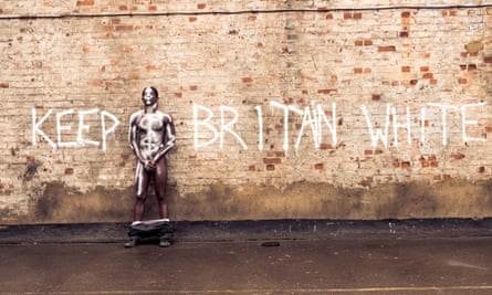 Young naked black male holding his crotch against a wall spray painted along with the words 'keep Britain white'
