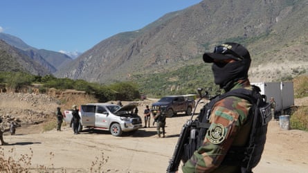 A man in army uniform holding a gun while a car is searched in the background