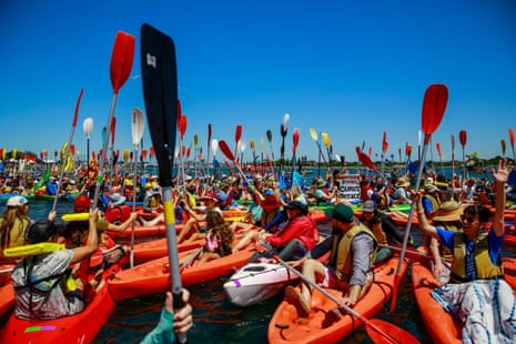 A sea of people in orange kayaks hold their paddles high in the air