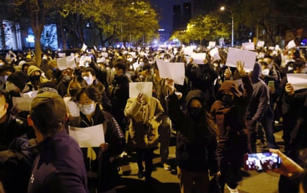 A large crowd of people wearing masks hold up blank pieces of paper