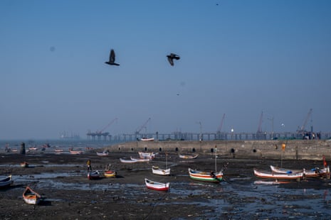 An expanse of shoreline at low tide with small fishing boats resting on a muddy beach and a highway on piles being built on the horizon