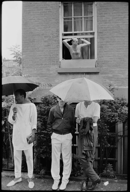 Men standing under umbrellas while another man stretches in the open window of a house behind them