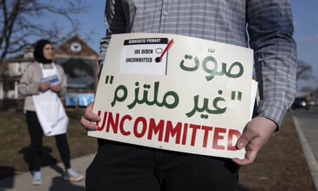 A man, pictured below the neck, holds a green and red sign in Arabic and English that says 'uncommitted,' beyond whom a woman in a headscarf stands on a curb.