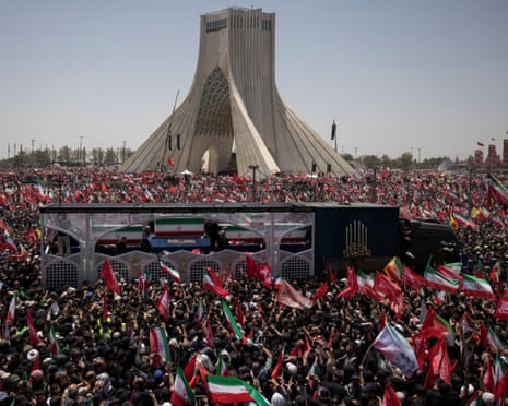 A truck moves through a crowd carrying Iranian flags in front of a building.
