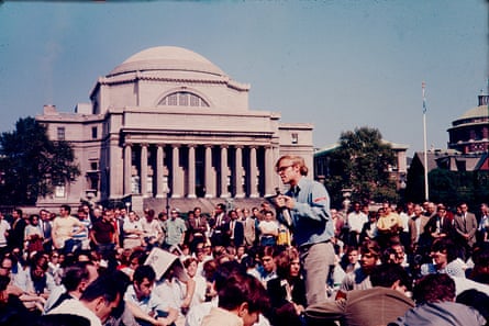 man stands with microphone as others sit on the ground around him, in front of columned and domed building, in a color photo