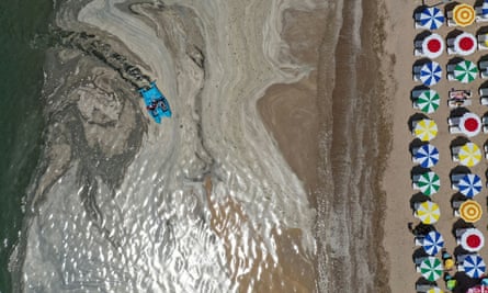Aerial view of a boat being paddled through a grey substance close to a beach with colourful umbrellas lined up in rows