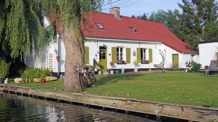 A low, white-washed house with green shutters and red roof sits under a willow directly adjacent to a canal